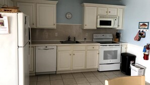 Una cocina con armarios blancos, microondas, horno, lavavajillas y un lavabo. Hay un reloj en la pared y un cubo de basura con etiqueta de reciclaje.