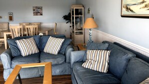 Una sala de estar con un sofá azul, una mesa de centro de madera y una lámpara. Se observa un comedor con una mesa y sillas al fondo.