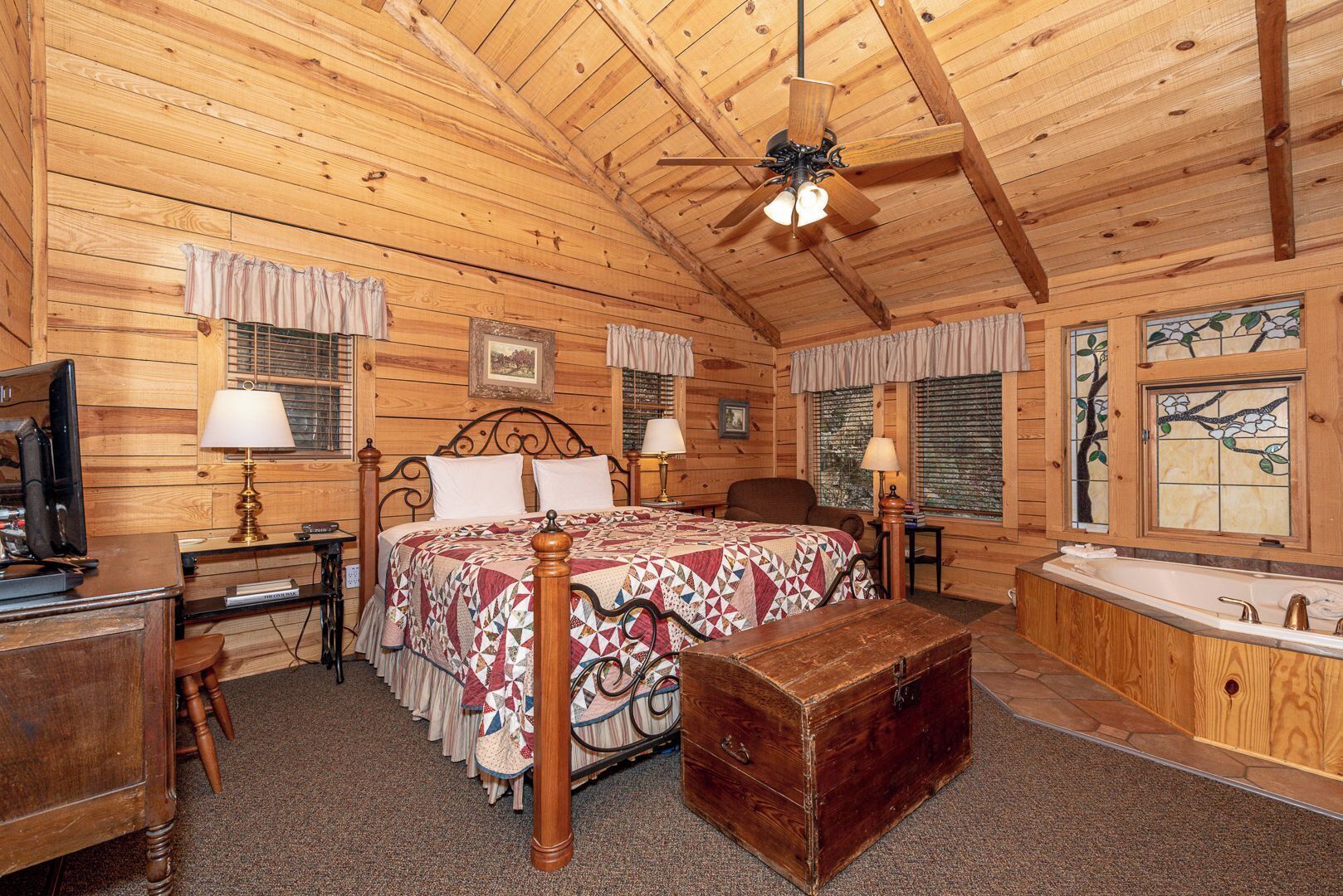 A wooden cabin bedroom with a bed, bedside tables, a desk, a dresser, a chest of drawers, a ceiling fan, and a large window with a view.