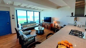 Ein geräumiges Wohnzimmer mit großem Fenster, einer Couch, einem Couchtisch und einer Küchenzeile inklusive Gaskochfeld und einer Obstschale.