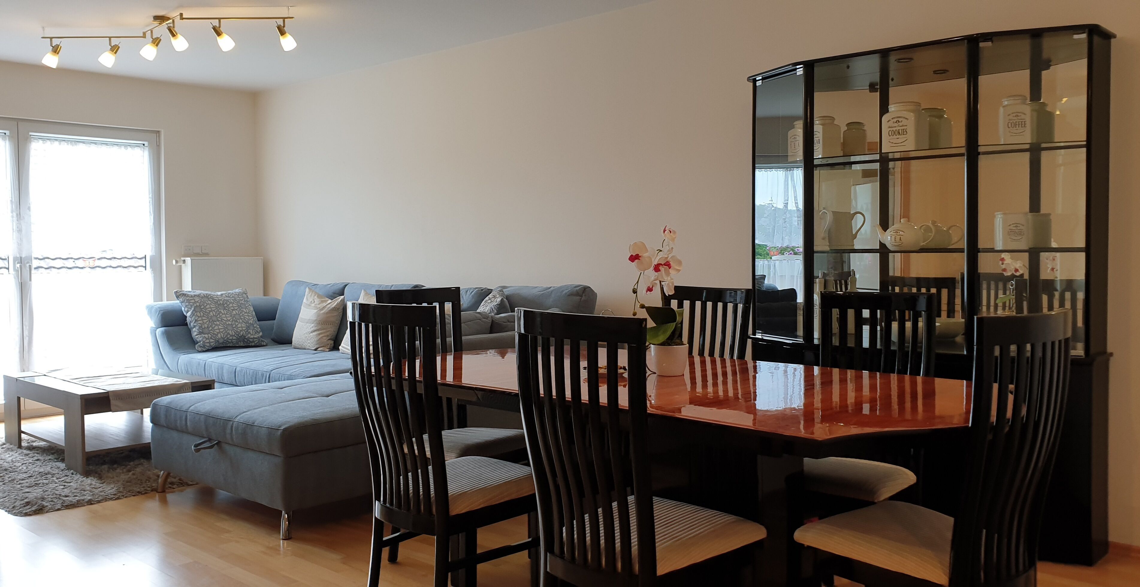 Uma sala de estar moderna com um sofá cinza, uma mesa de jantar de madeira com cadeiras, um armário de vidro com utensílios de cozinha e uma pequena planta em vaso.