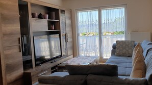 A living room with a sofa, TV, wooden cabinet, and a balcony with a view. - Ferienwohnung Maria - Bad Kissingen (Bad Kissingen)