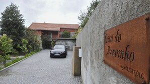 Un carrer asfaltat que condueix a un edifici amb teules vermelles. Un cotxe està estacionat al carrer. Un cartell a la paret diu "Casa do Azevinho" amb un número d'identificació.