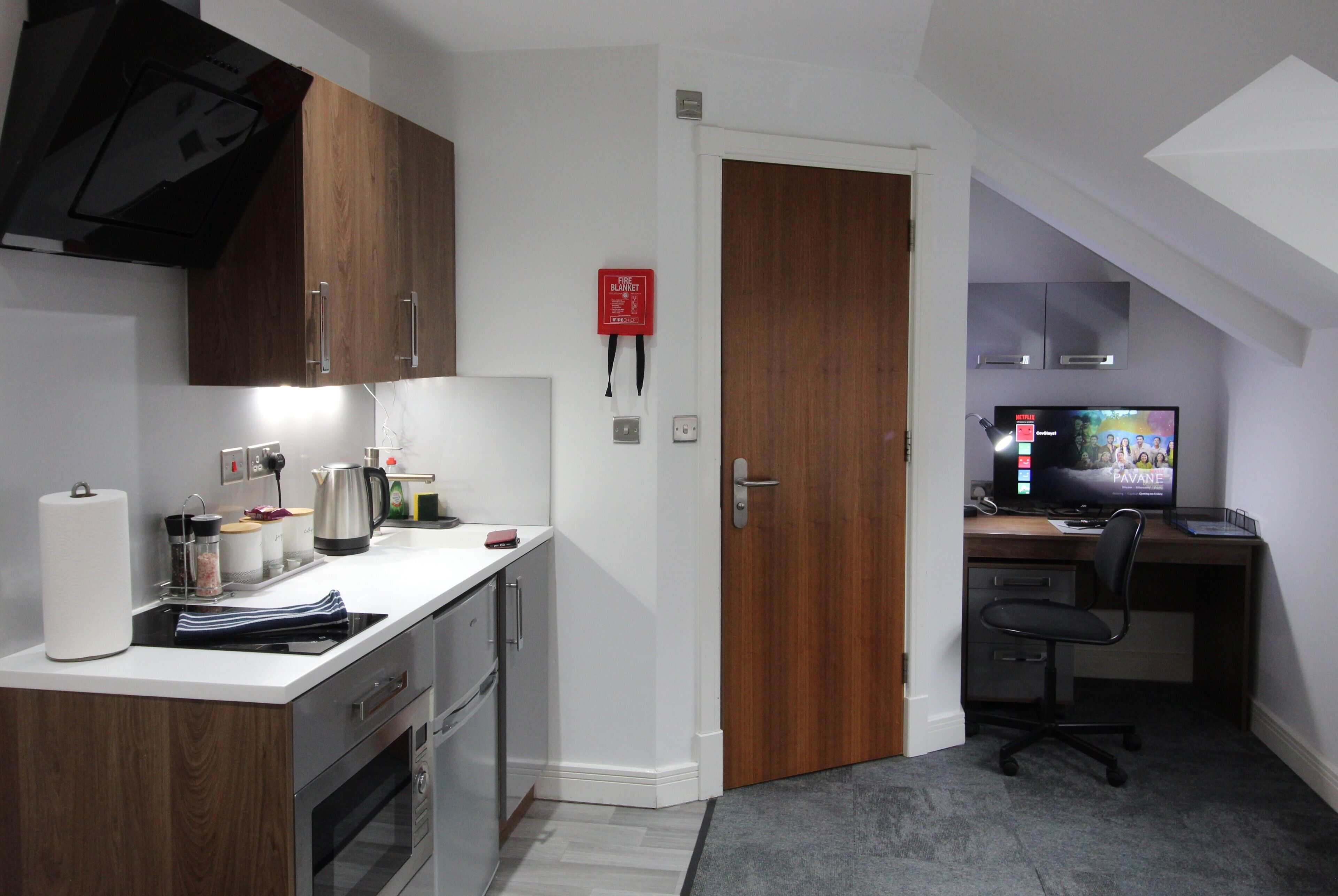 A compact kitchen with a stove, oven, and a coffee maker. There is a wooden door leading to a bedroom with a desk and a computer monitor.