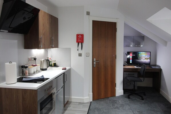 A compact kitchen with a stove, oven, and a coffee maker. There is a wooden door leading to a bedroom with a desk and a computer monitor.