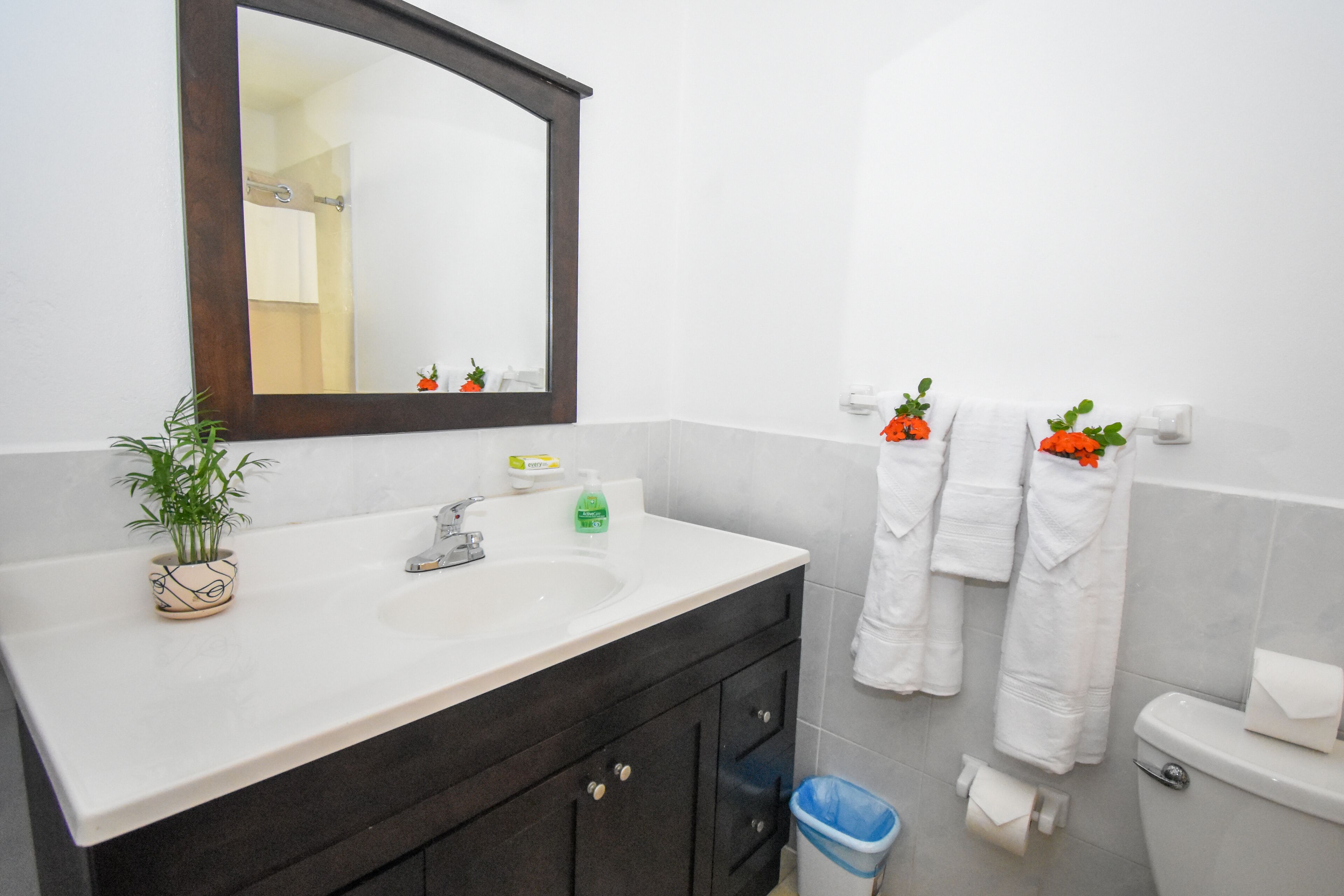 Un baño con un lavamanos blanco, armarios oscuros, un espejo, toallas blancas y un cubo de basura azul.