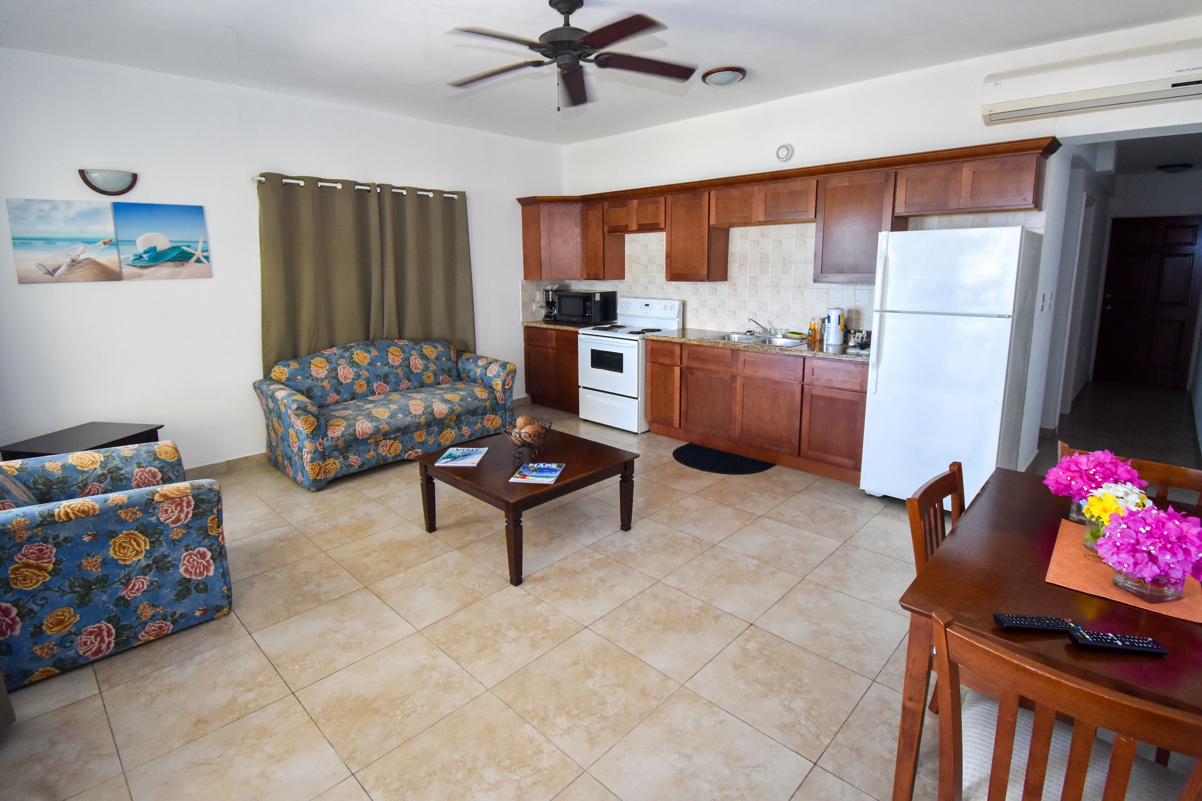 A living room with a sofa, coffee table, and a dining table with chairs. There is a kitchen area with wooden cabinets, a refrigerator, and a microwave.