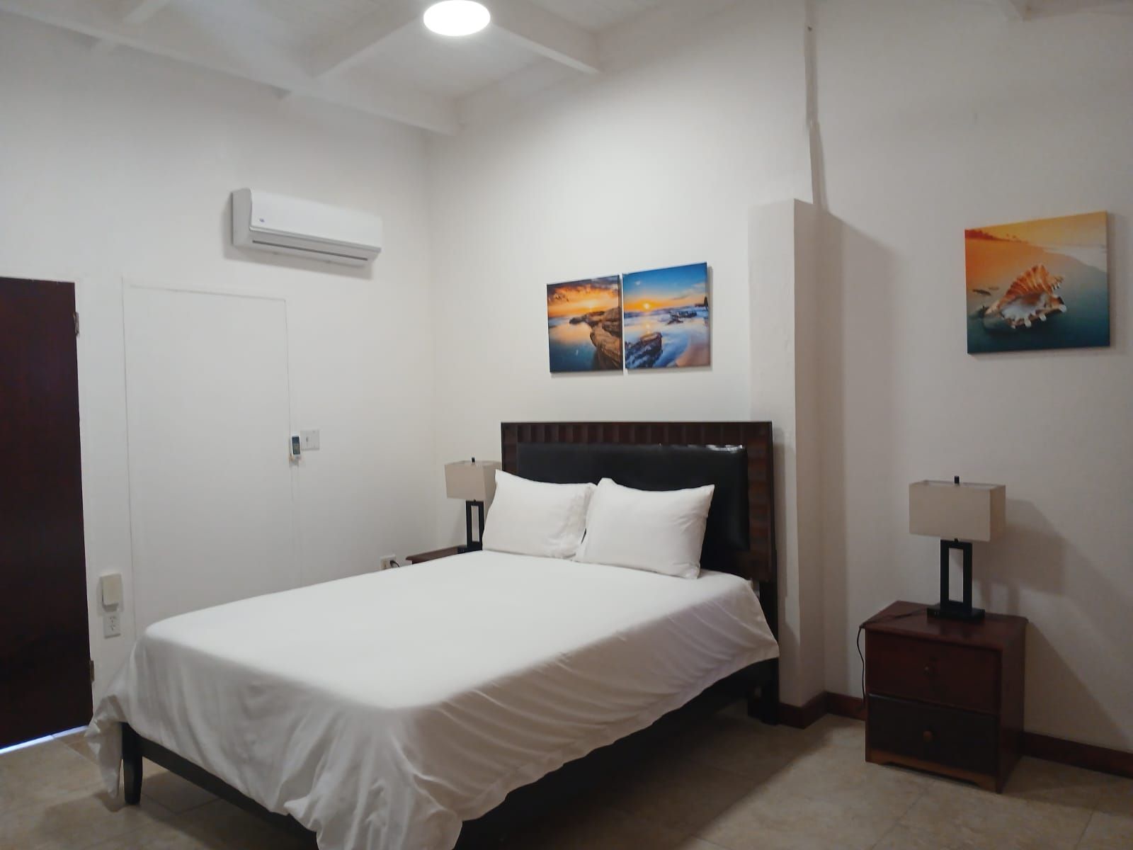Une chambre d’hôtel comprenant un lit, des lampes de chevet, une table de nuit, un climatiseur fixé au mur et deux tableaux.