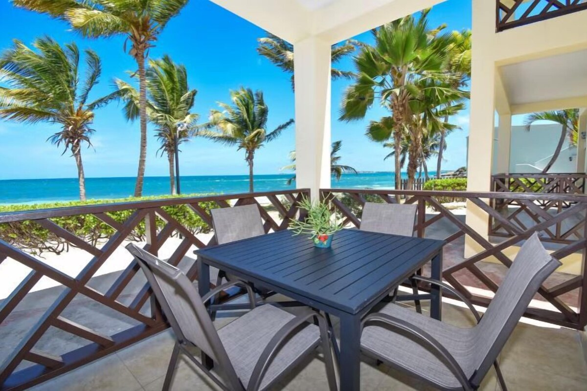 Un balcón con mesa y sillas, con vista a una playa con palmeras y el océano.