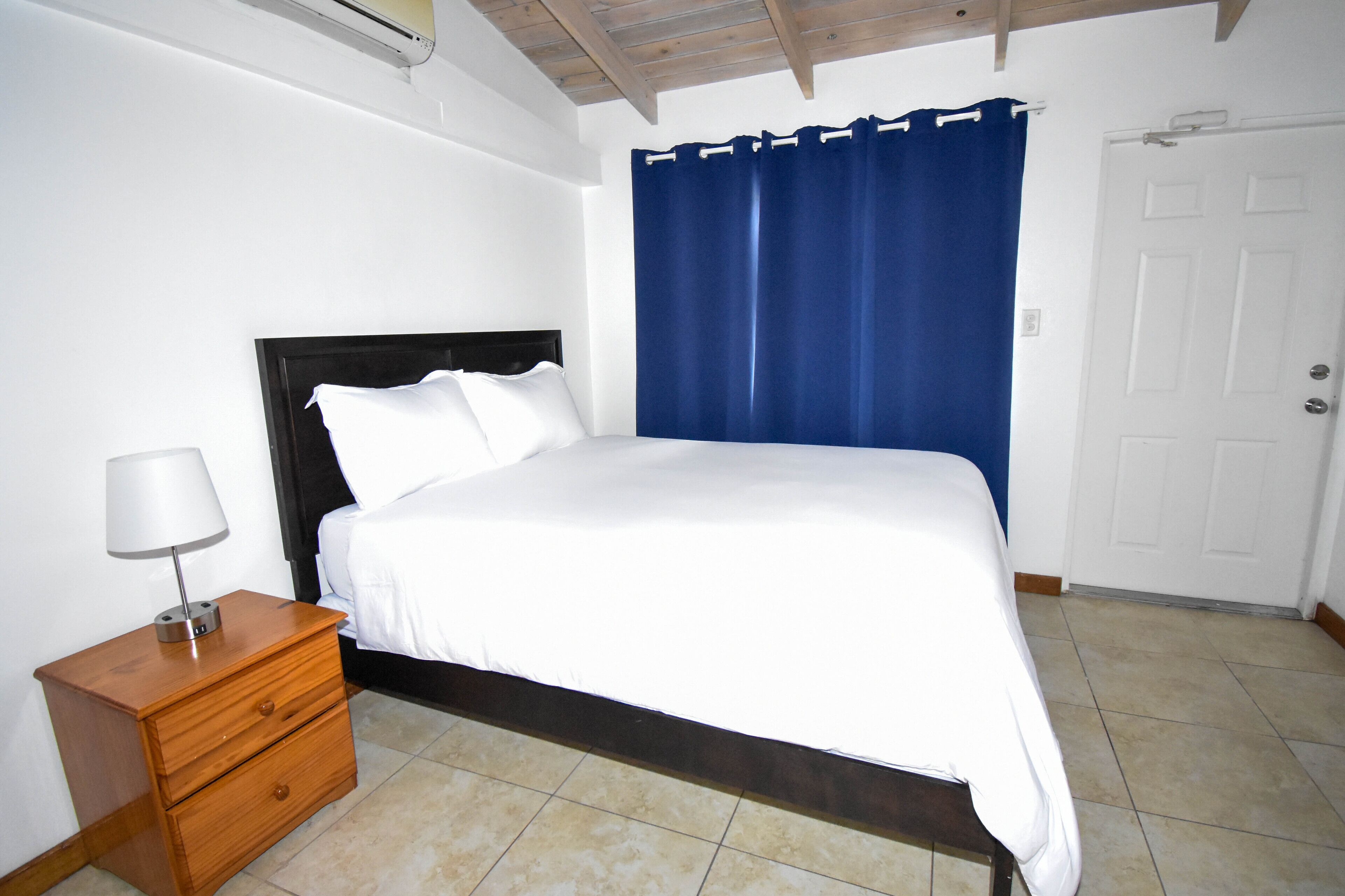 A bedroom with a bed, a nightstand, a lamp, a blue curtain, and a white door.