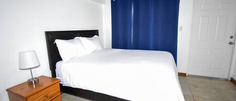 A bedroom with a bed, a nightstand, a lamp, a blue curtain, and a white door.