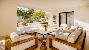 A modern living room with a sofa, coffee table, and chairs, featuring a view of palm trees and a pool. - Oceanfront Luxury Escape, Breathtaking Views. Private Pool & Jacuzzi (Cabo San Lucas)