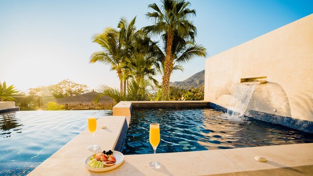 A poolside setting with a clear view of the pool, a water feature, and a plate of fruit and a glass of orange juice. - Oceanfront Luxury Escape, Breathtaking Views. Private Pool & Jacuzzi (Cabo San Lucas)