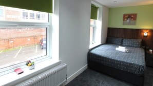 A bedroom with a large window, a radiator, a bed with a patterned bedspread, and a small plant on the windowsill. - Victoria House - Classic Studios by CovStays (Coventry)