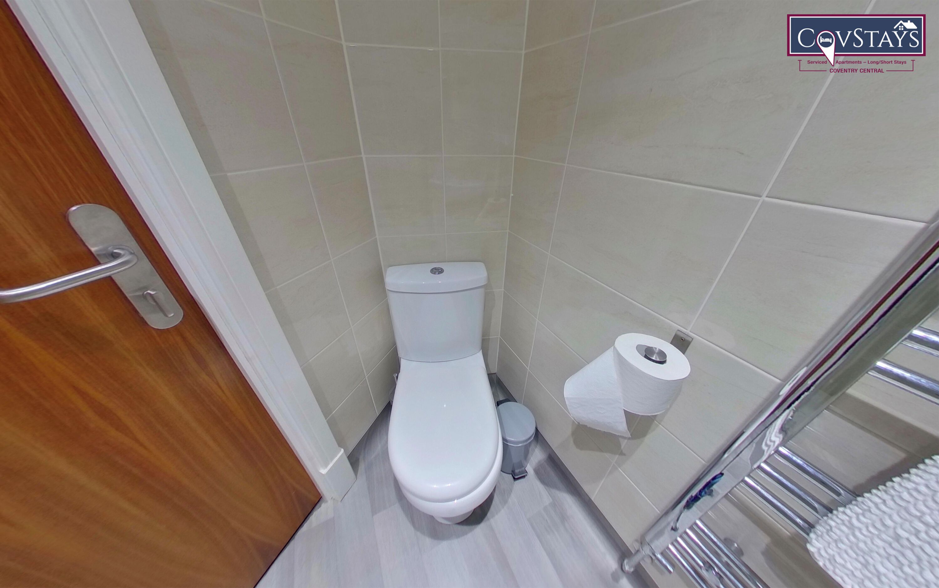 Un baño pequeño con azulejos, un inodoro, un rollo de papel higiénico y un perchero de metal.