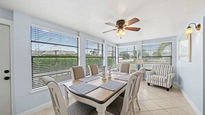Comedor con una mesa dispuesta para cuatro personas, sillas alrededor, un ventilador de techo y ventanas amplias con persianas.
