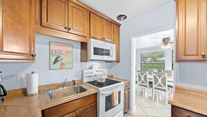 Cocina con armarios de madera, microondas, estufa, lavabo y comedor con sillas blancas.
