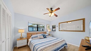 Un dormitorio con una cama, ventilador de techo, ventana, mesita de noche, lámpara, espejo y escritorio de madera.