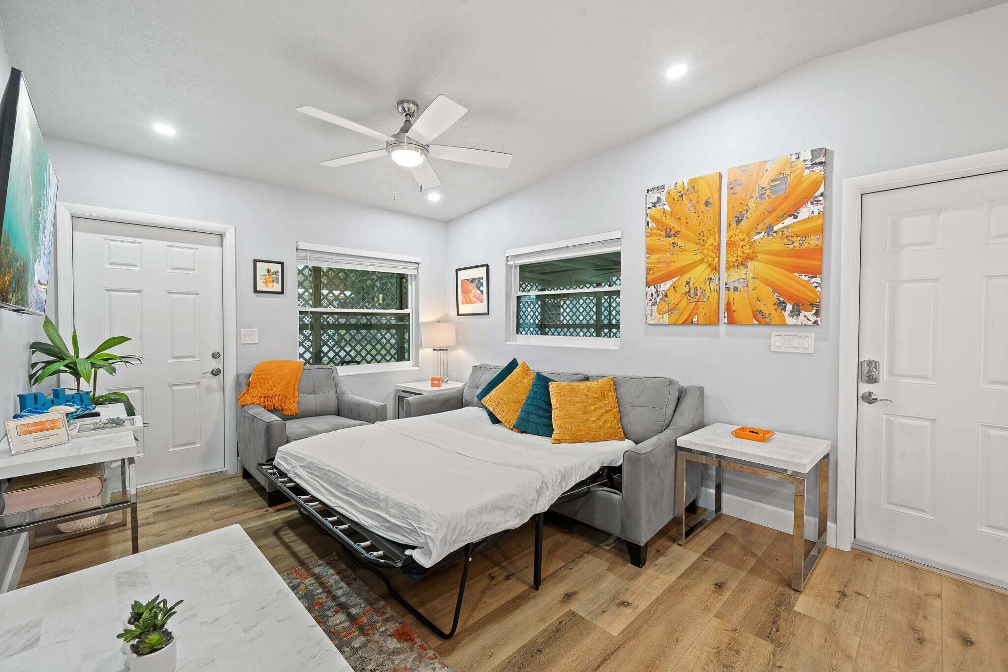 A modern bedroom with a bed, a desk, and a chair. There is a ceiling fan, a window with a view, and a door.
