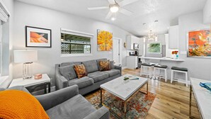 A modern living room with a gray sofa, a coffee table, and a ceiling fan. There is a kitchen area with white cabinets and a dining table. - Midtown Cottage - Walk to it All! (Cocoa Beach)