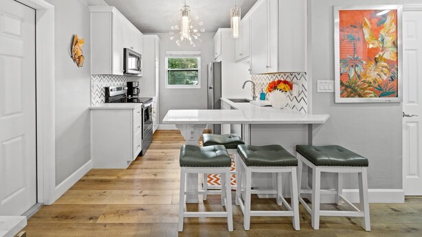 A modern kitchen with white cabinetry, a central island with stools, a microwave, and a hanging light fixture. - Midtown Cottage - Walk to it All! (Cocoa Beach)
