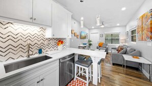 A modern kitchen with white cabinets and a herringbone tile backsplash. There is a dining area with a gray sofa and a coffee table. - Midtown Cottage - Walk to it All! (Cocoa Beach)