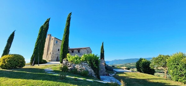 Un bâtiment en pierre doté d’une tour, entouré d’une végétation bien entretenue et surplombé d’un ciel bleu clair.