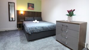 A bedroom with a bed, a dresser, a nightstand, a mirror, and a wall-mounted lamp.