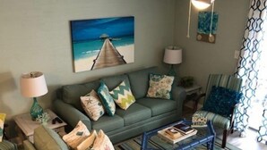 A living room with a green sofa, patterned cushions, a coffee table, a painting of a beach, a ceiling fan, and a patterned chair. - NEED SPACE? Entire Townhouse-Large 2-Bdrm 1,150sf Steps to Beach. 5% Off 7Nights (Myrtle Beach)