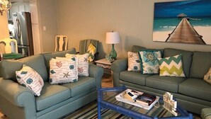 A living room with two green sofas, a glass coffee table, and a painting of a beach scene. - NEED SPACE? Entire Townhouse-Large 2-Bdrm 1,150sf Steps to Beach. 5% Off 7Nights (Myrtle Beach)