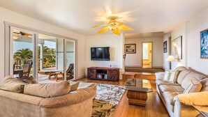 A spacious living room with a ceiling fan, a flat-screen TV, and a large window offering a view of palm trees. - Coastline View (Kailua-Kona)