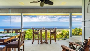 A dining area with a table and chairs, large windows with a view of the ocean, and a ceiling fan. - Coastline View (Kailua-Kona)