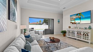 A spacious living room with a white sofa, a round coffee table, and a TV displaying a beach scene. - LimitedTime Special Offer  New Vanderbilt Beach Home Heated Saltwater Pool & Spa (Naples)