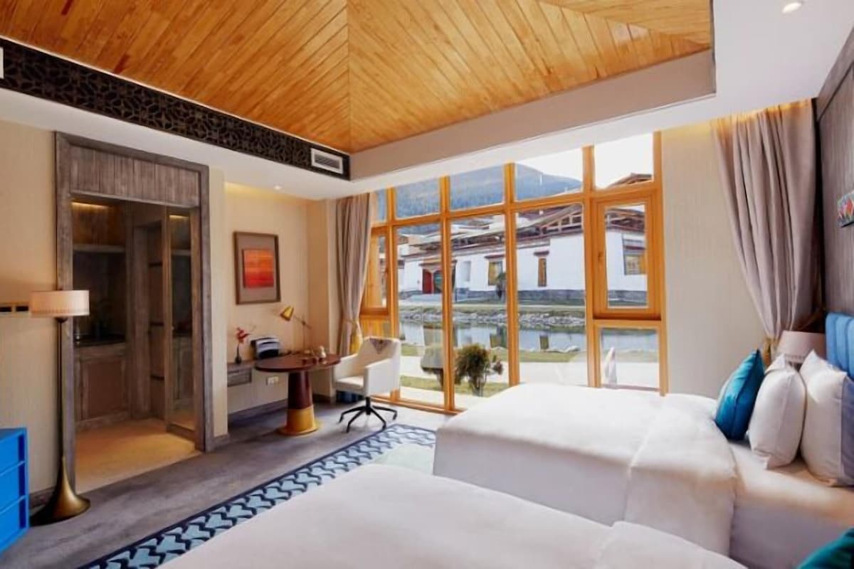 A hotel room with a wooden ceiling, large windows offering a view of buildings and a body of water, a desk with a chair, and two beds.