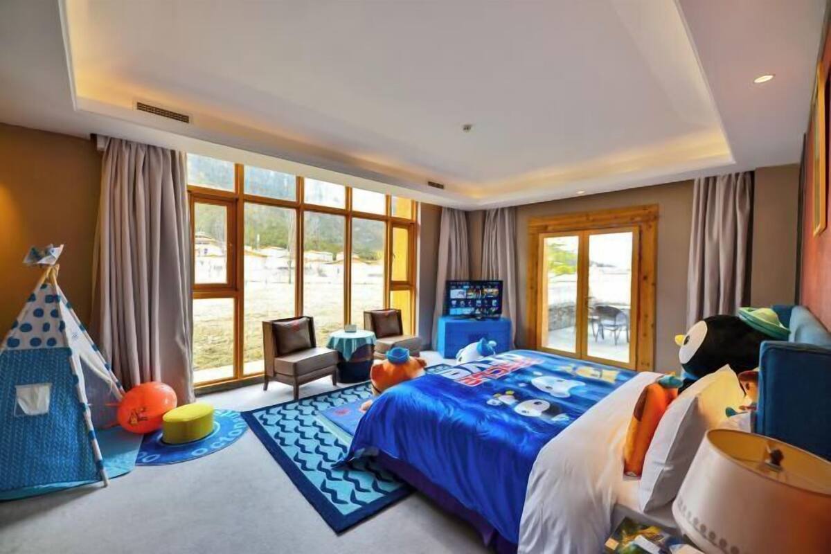 A hotel room with a large bed, a play area with a tent and toys, a TV, and large windows offering a view of the outdoors.
