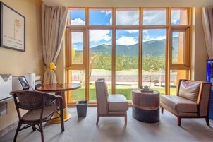 Habitación de hotel con un ventanal grande que ofrece vistas a la montaña, una mesa redonda, dos sillas, un sofá y un bote de basura pequeño.