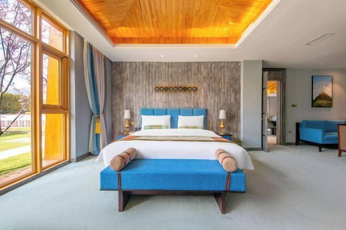 A large bed with a blue headboard, a wooden ceiling, and a view of the outdoors through large windows.