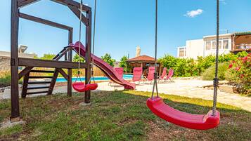 A playground with swings and a slide, a pool, and a gazebo in a backyard.