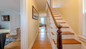 A wooden staircase with a handrail leading to a second-floor landing. A door on the left leads to a bedroom with a bed and a chair. - Classic Mystic Retreat Walking Distance to Downtown (Groton)