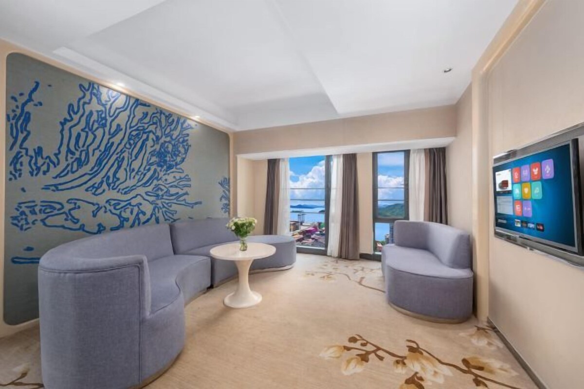 A modern hotel room with a curved grey sofa, a small white table, and a large TV screen.