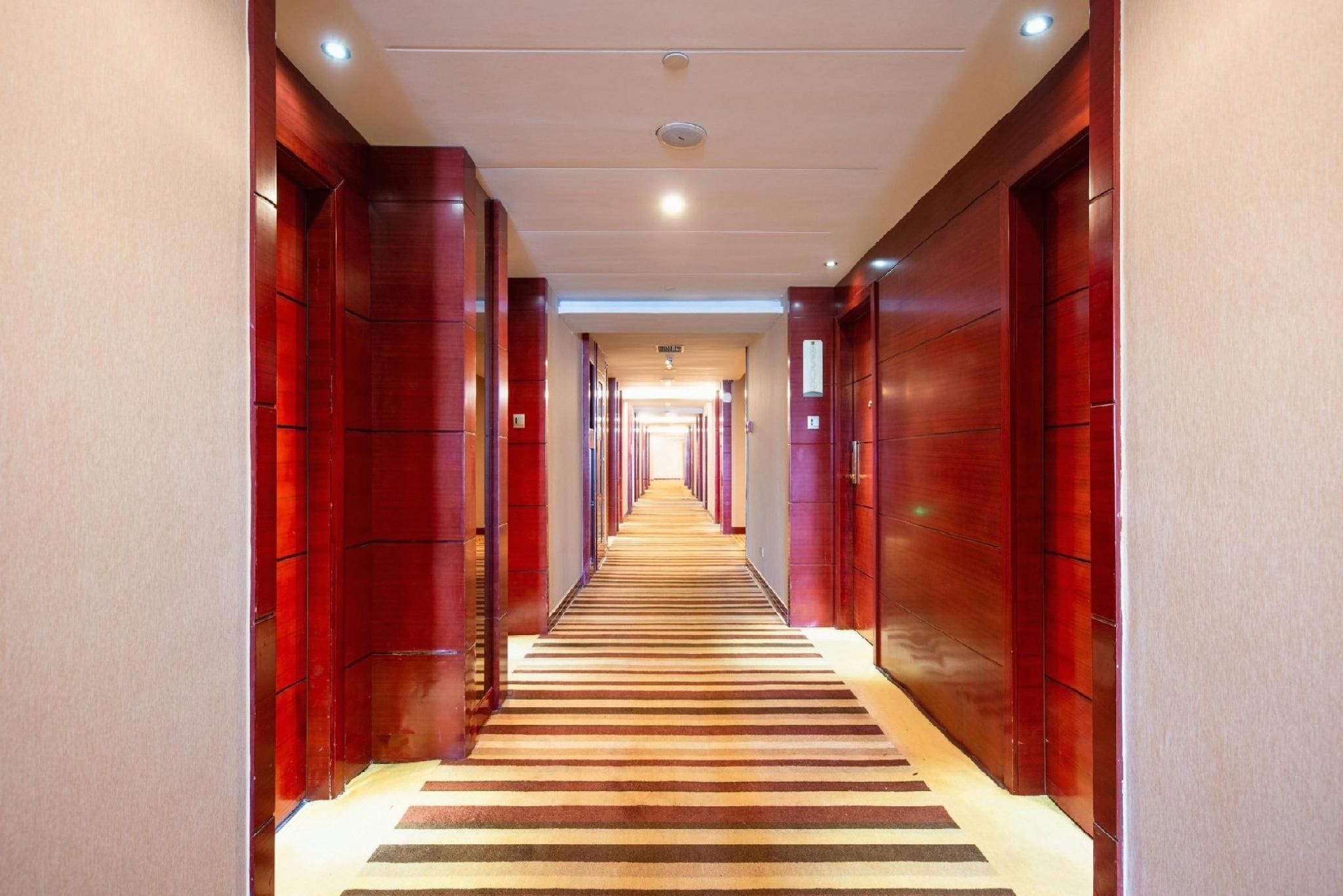 Un pasillo de hotel con puertas de madera, alfombra a rayas e iluminación empotrada en el techo.
