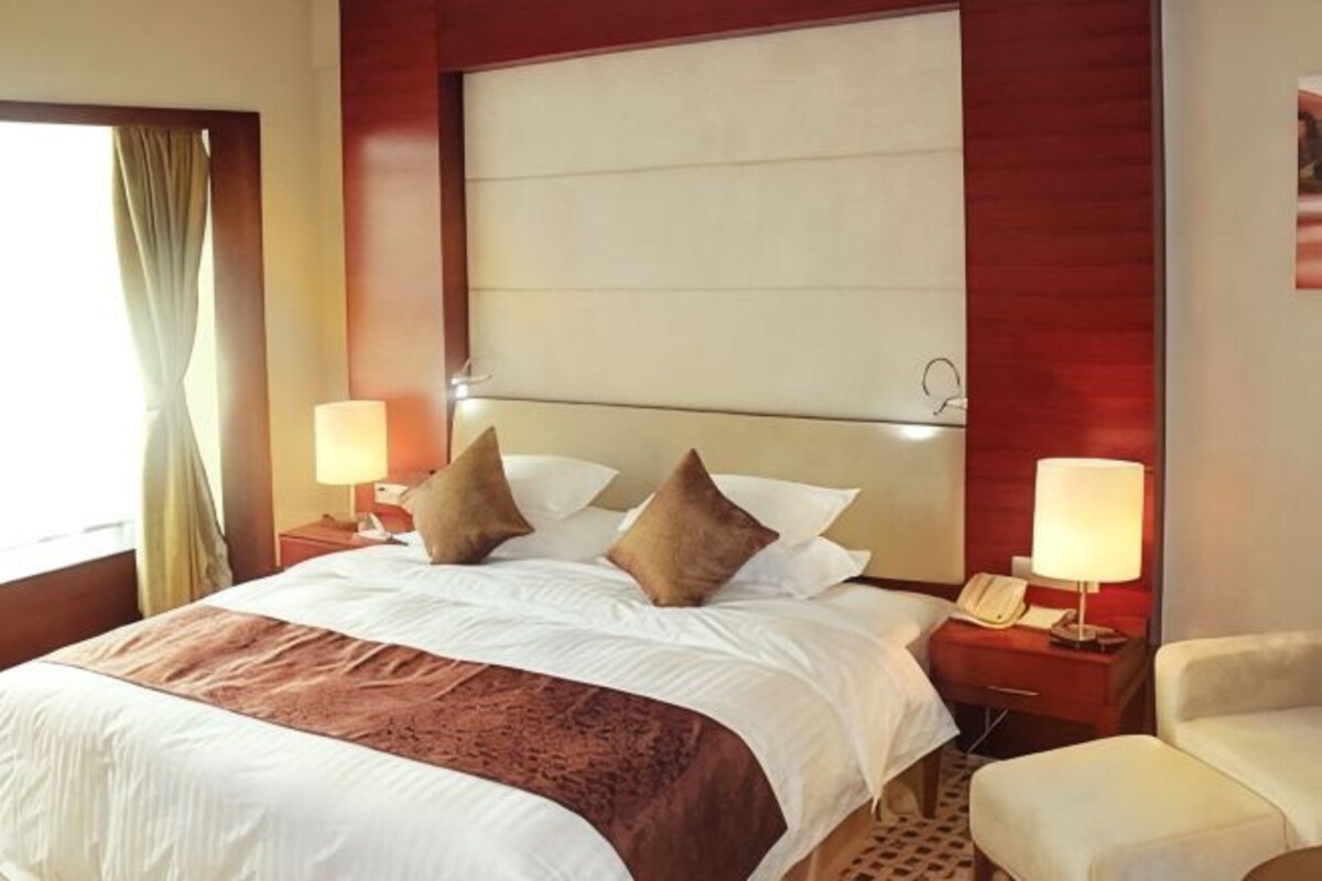 Habitación de hotel con una cama grande, dos mesitas de noche con lámparas, una silla y un ventanal con cortinas.