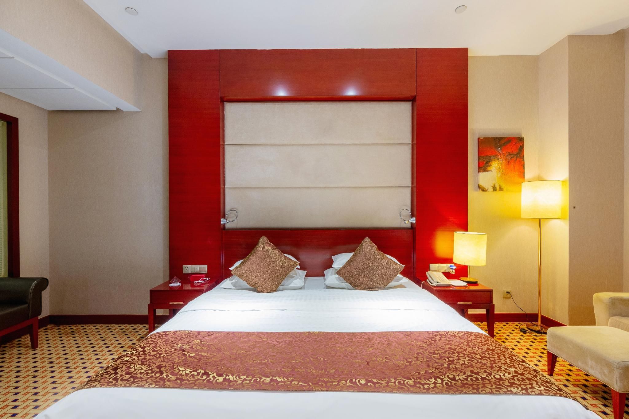 A hotel room with a large bed, a red headboard, a bedside table with a lamp, a chair, and a patterned carpet.