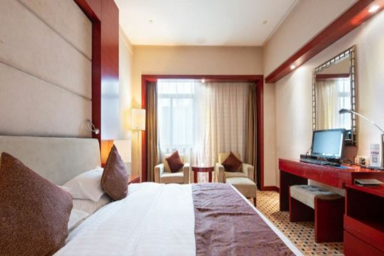 Habitación de hotel con cama, escritorio con computadora, dos sillas, ventana con cortinas y un espejo en la pared.