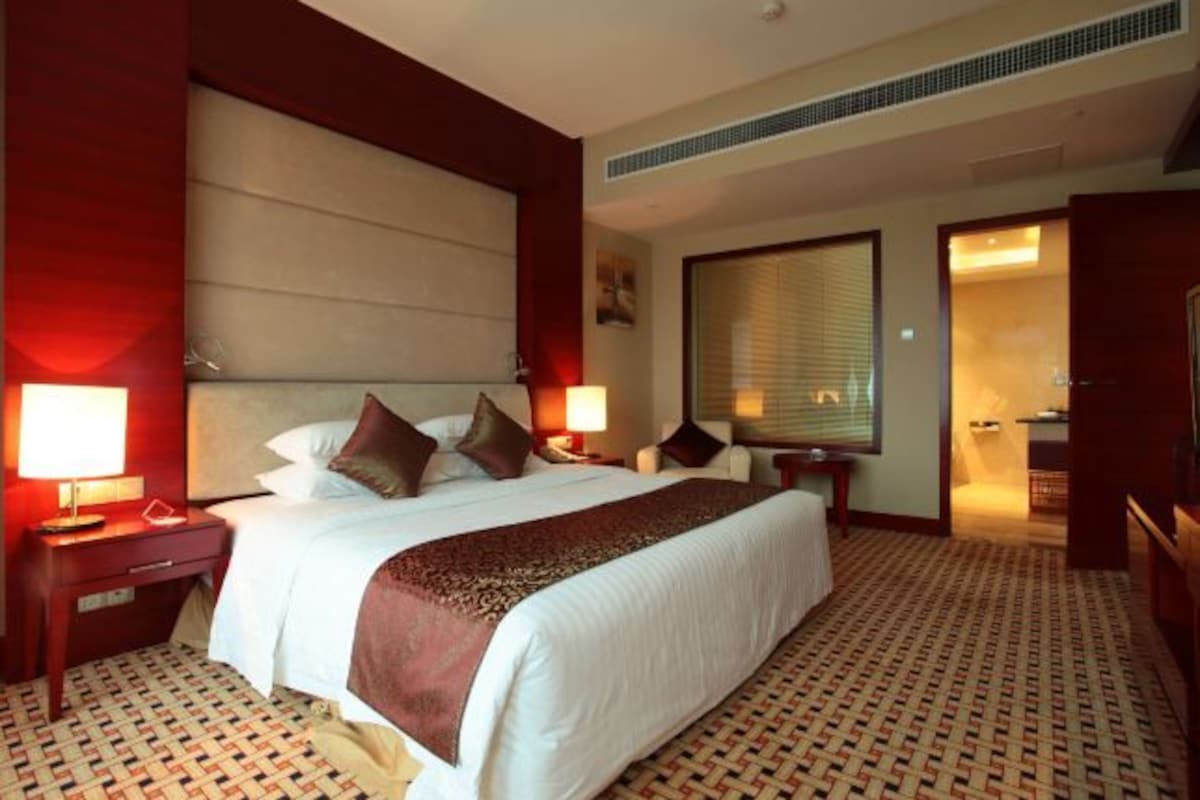 Habitación de hotel con una cama grande, dos mesitas de noche con lámparas, un ventanal con persianas y una alfombra estampada.