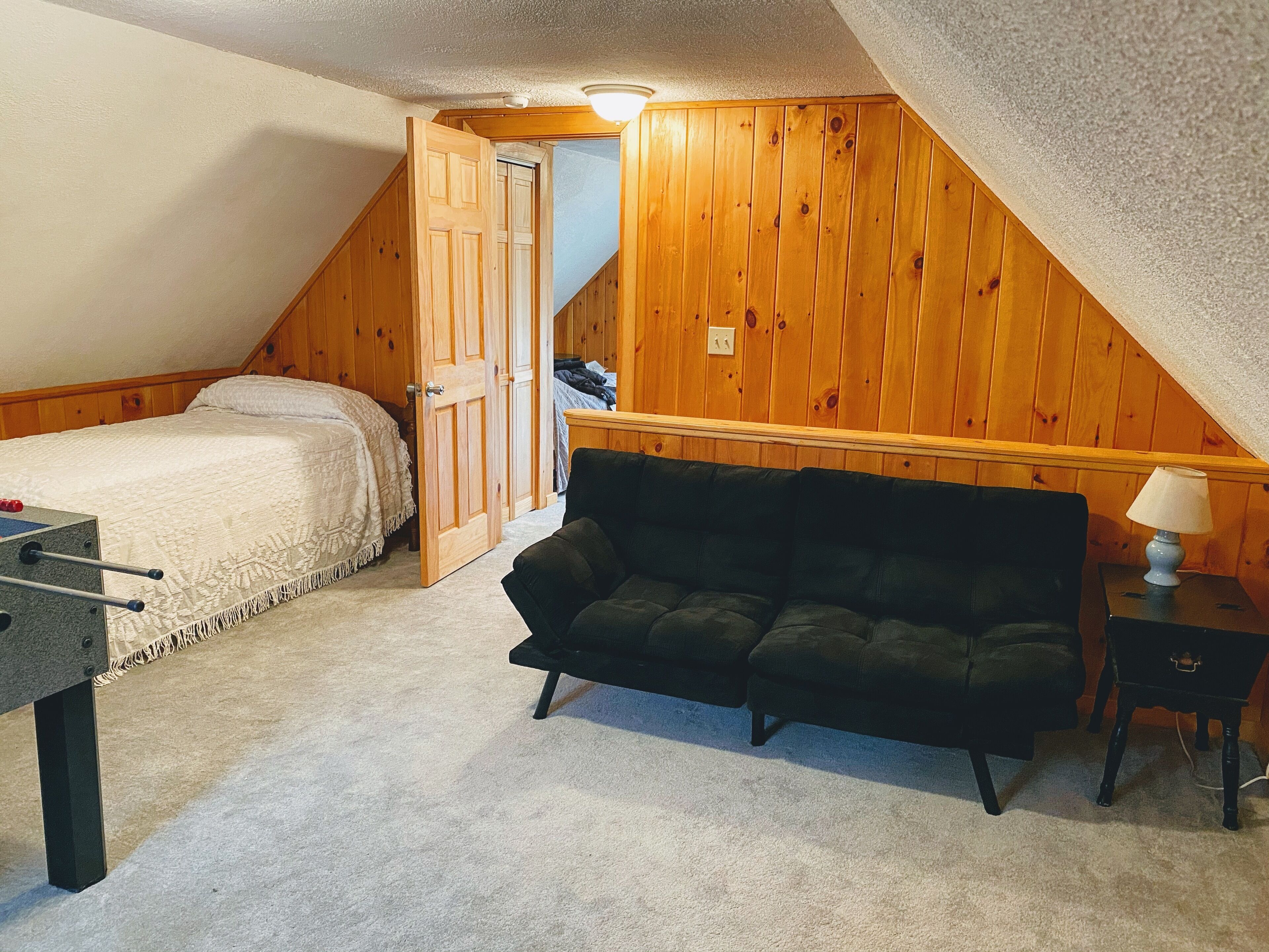 A bedroom with a bed, a dark sofa, a small table with a lamp, and a wooden wall.