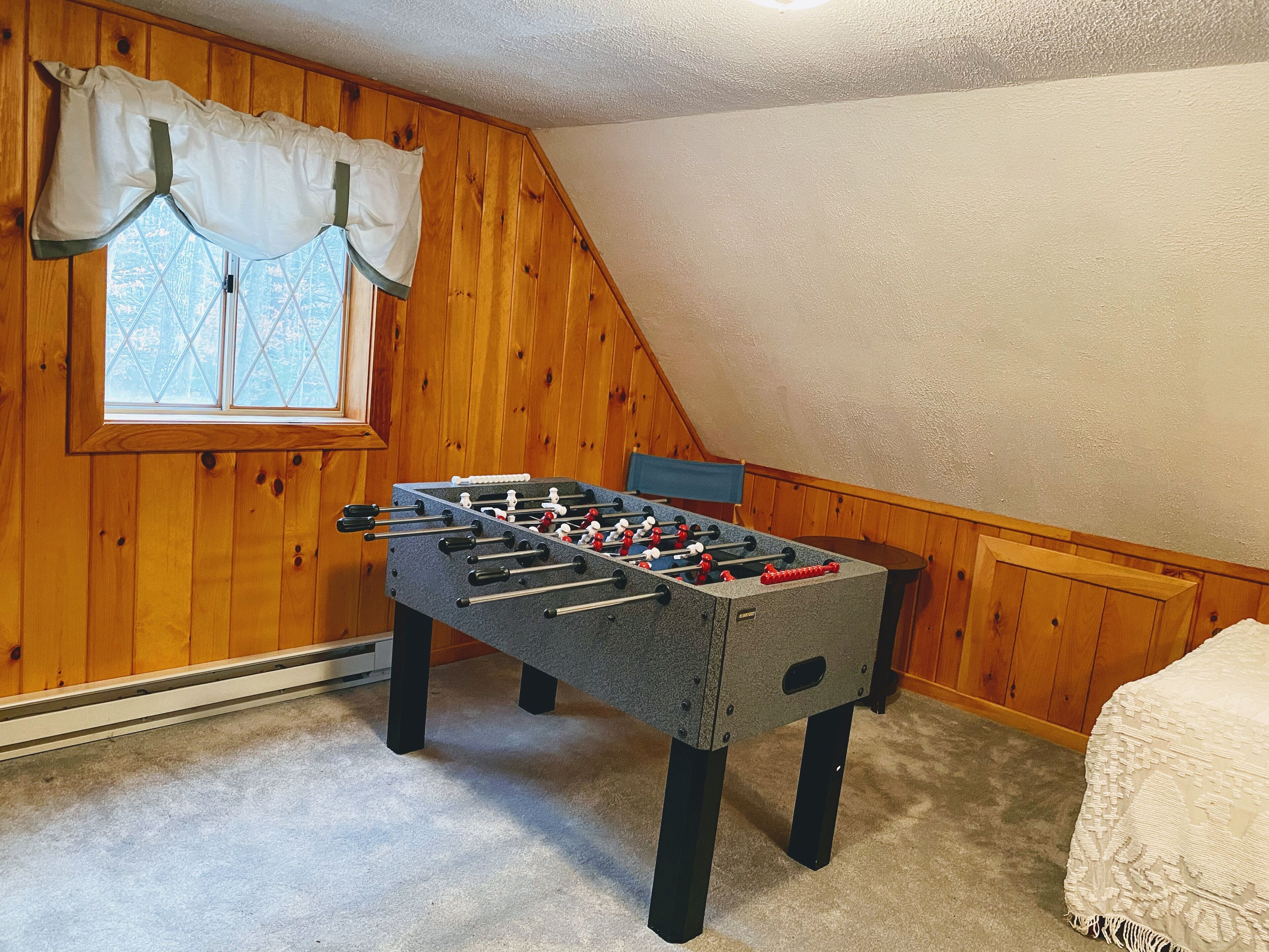Una habitación con una mesa de fútbol de mesa, paredes de madera, una ventana con cortinas y una cama con cubrecolcha blanco.