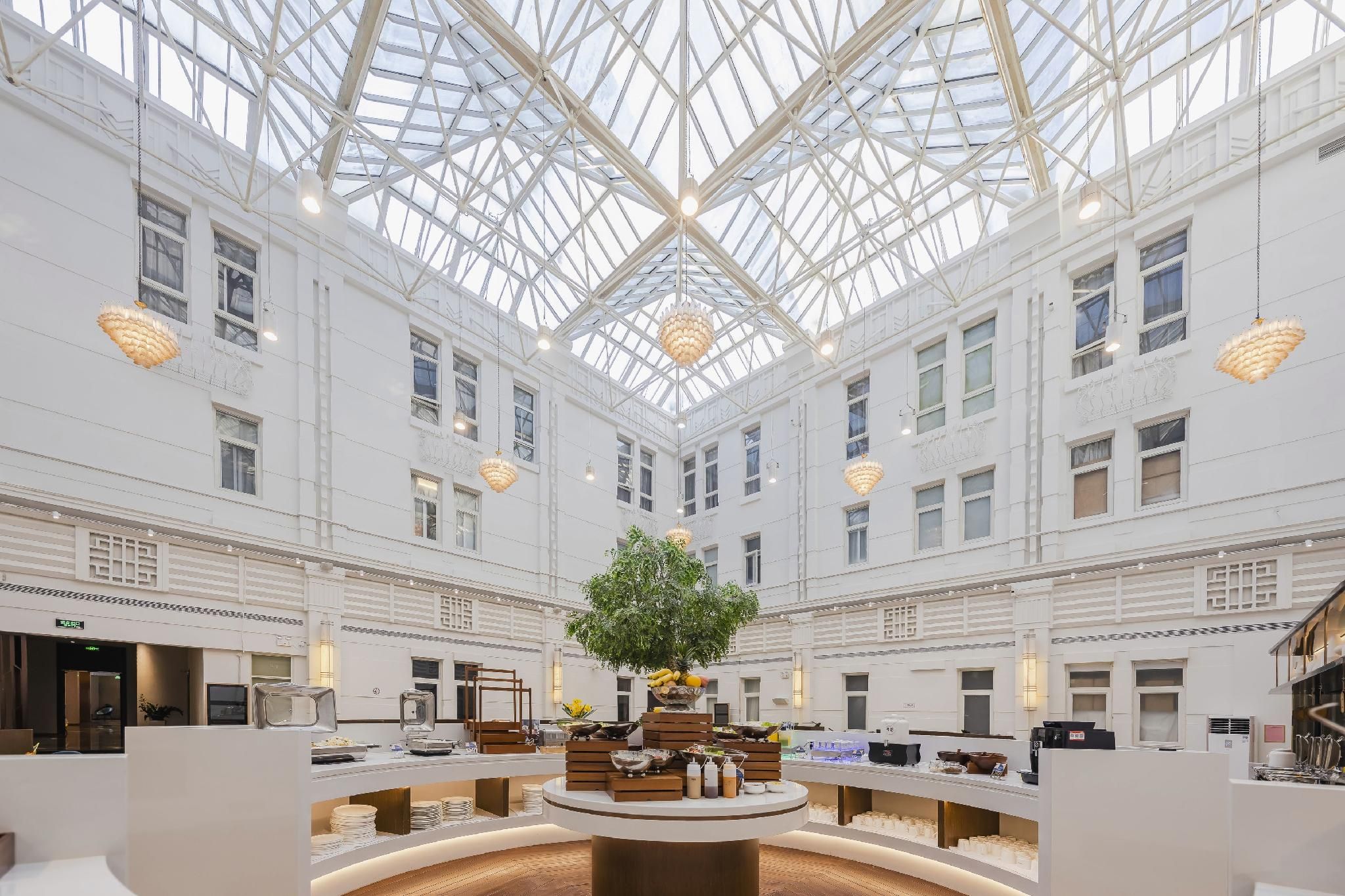 A spacious atrium with a high glass ceiling, a central tree, and a variety of amenities including a coffee station and seating areas.