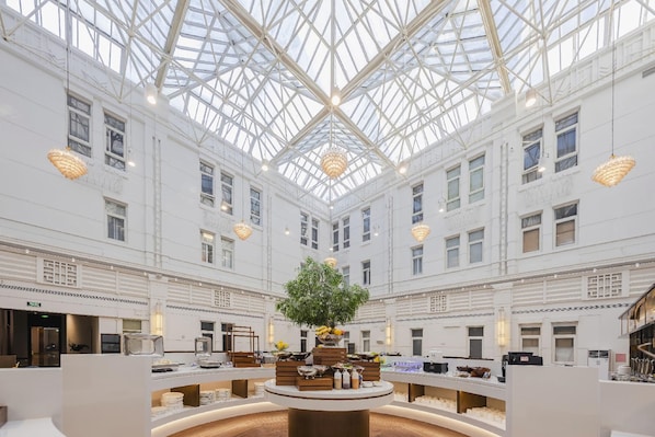 A spacious atrium with a high glass ceiling, a central tree, and a variety of amenities including a coffee station and seating areas. - Yitel (Shanghai)