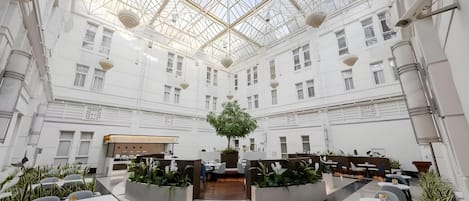 A spacious atrium with a high ceiling and a central tree, surrounded by seating areas and a reception desk.
